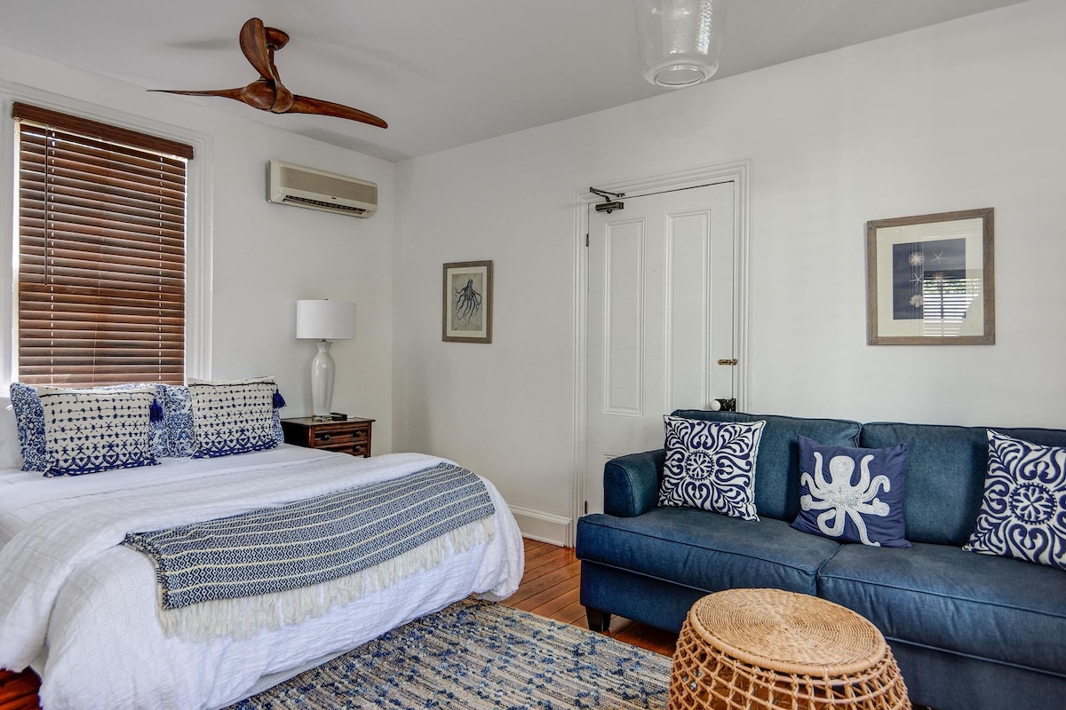 The living/bedroom area features a spacious bed adorned with patterned pillows, accompanied by a stylish sofa in deep blue. Artwork is displayed on white walls, while a woven rug adds texture to the hardwood floor. Natural light filters through wood-blind covered windows.