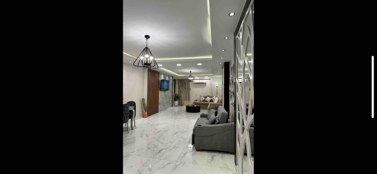 An open living area features a marble floor and modern furnishings, including a dark gray sofa and accent chairs. A television is mounted on the wall, with a decorative plant in the corner. Soft lighting enhances the contemporary design.