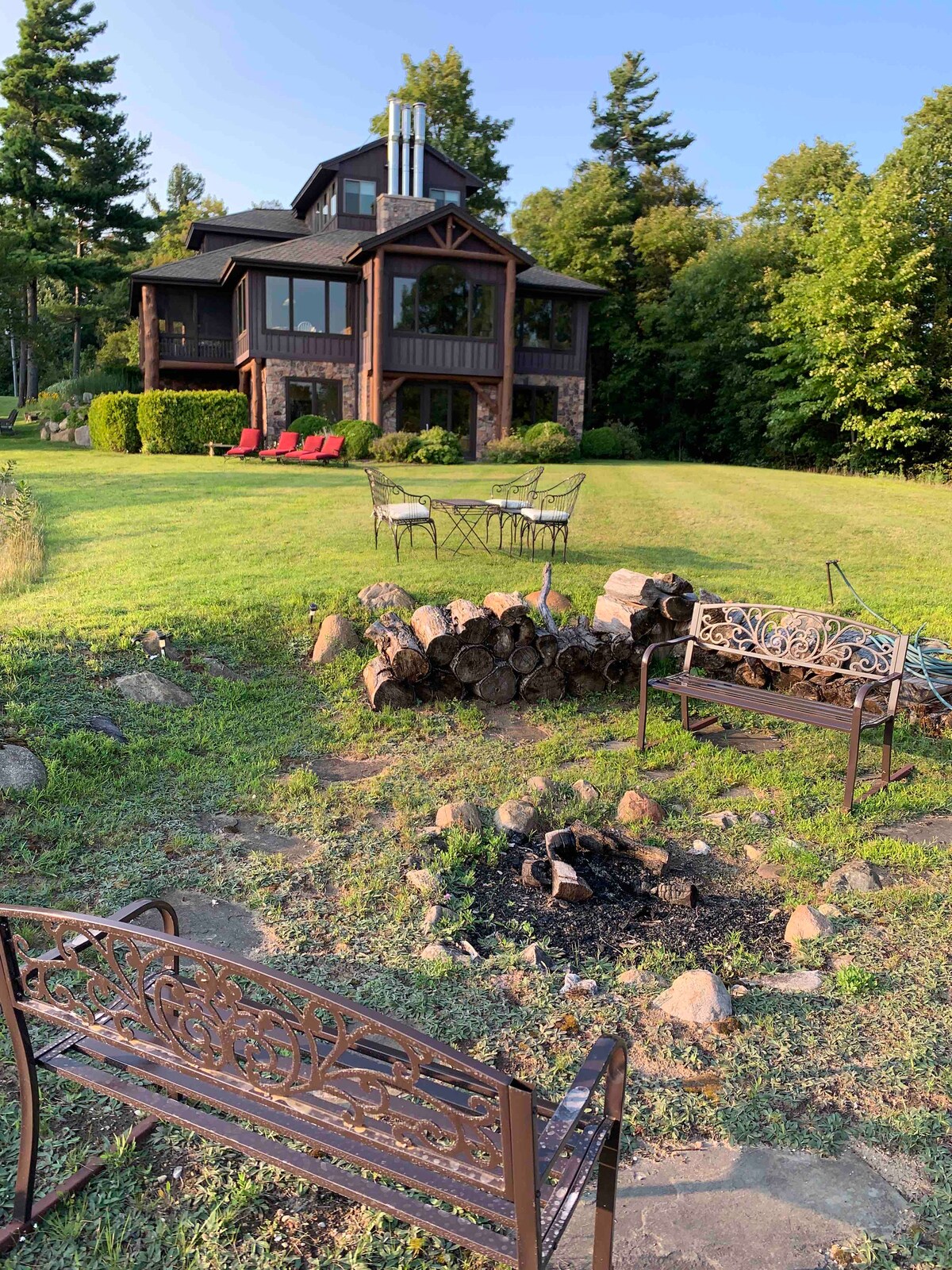 A spacious backyard area features a fire pit surrounded by seating options. A stack of firewood is nearby, while the home with large windows and a distinctive design stands in the background, framed by lush greenery.