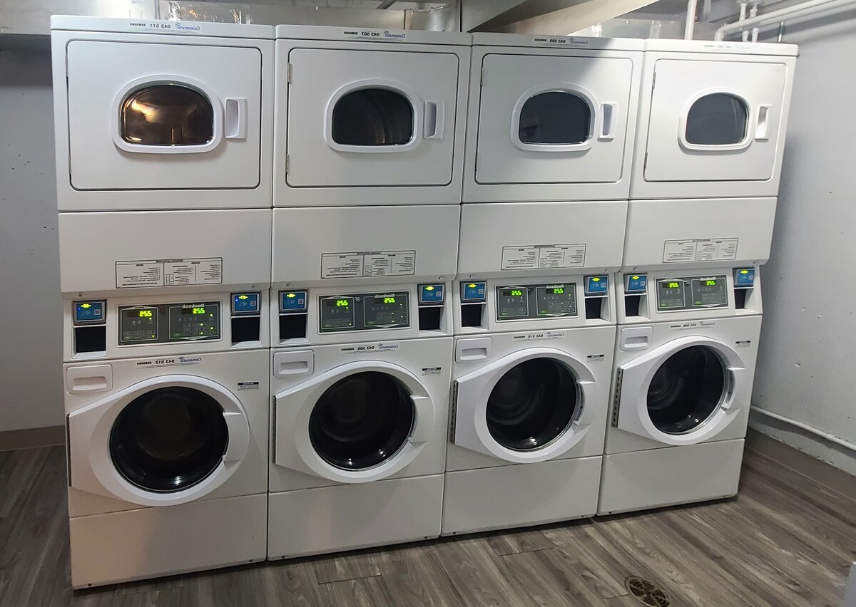 A row of stacked washer and dryer units is shown in a clean, well-lit laundry area. The machines feature digital displays and are positioned against a plain wall, with bright overhead lighting illuminating the space.