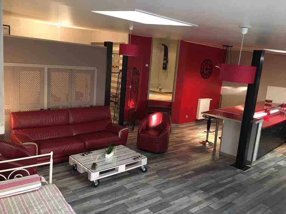 The spacious living area features a red leather sofa, a matching armchair, and a glass coffee table. Natural light filters through a skylight, illuminating the open layout that connects the living space to the kitchen area in the background.