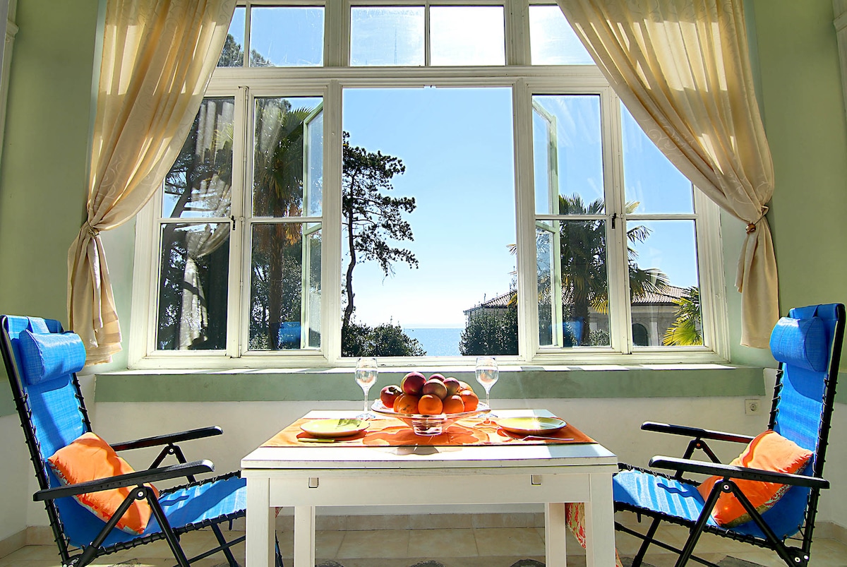A bright dining area is featured, showcasing a small table set for two with colorful plates and a centerpiece of fresh fruit. Large windows allow natural light to fill the space, offering serene views of the sea and surrounding trees.