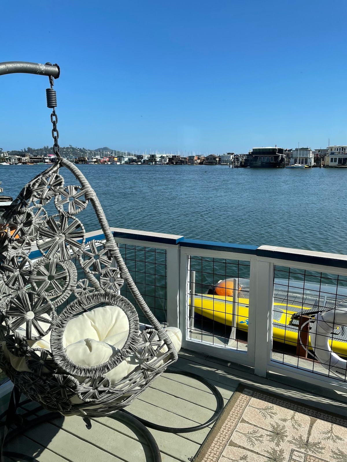 A hanging chair made of woven material is positioned on the deck, overlooking the water. Calm blue water stretches out, with colorful kayaks visible nearby. In the background, floating homes line the lagoon. The scene is complemented by clear skies and distant hills.