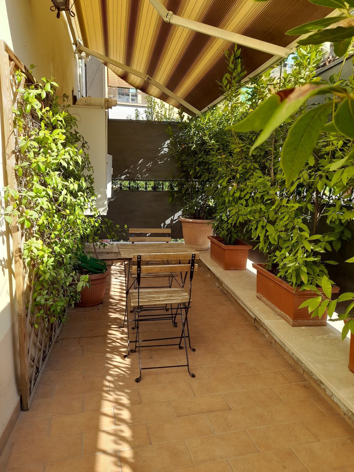 A tranquil terrace is surrounded by lush greenery, featuring potted plants and a small wooden table with two folding chairs. Sunlight filters through the awning, casting soft shadows on the tiled floor. The serene outdoor space invites moments of relaxation.