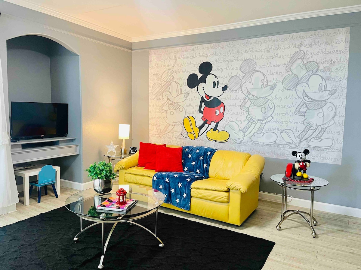 The living room features a Mickey Mouse mural, complemented by a yellow sofa adorned with a blue star-patterned blanket. A glass coffee table sits in the center, while a small blue chair and a television are positioned against a light gray wall. Decorative items include a plush Mickey Mouse.
