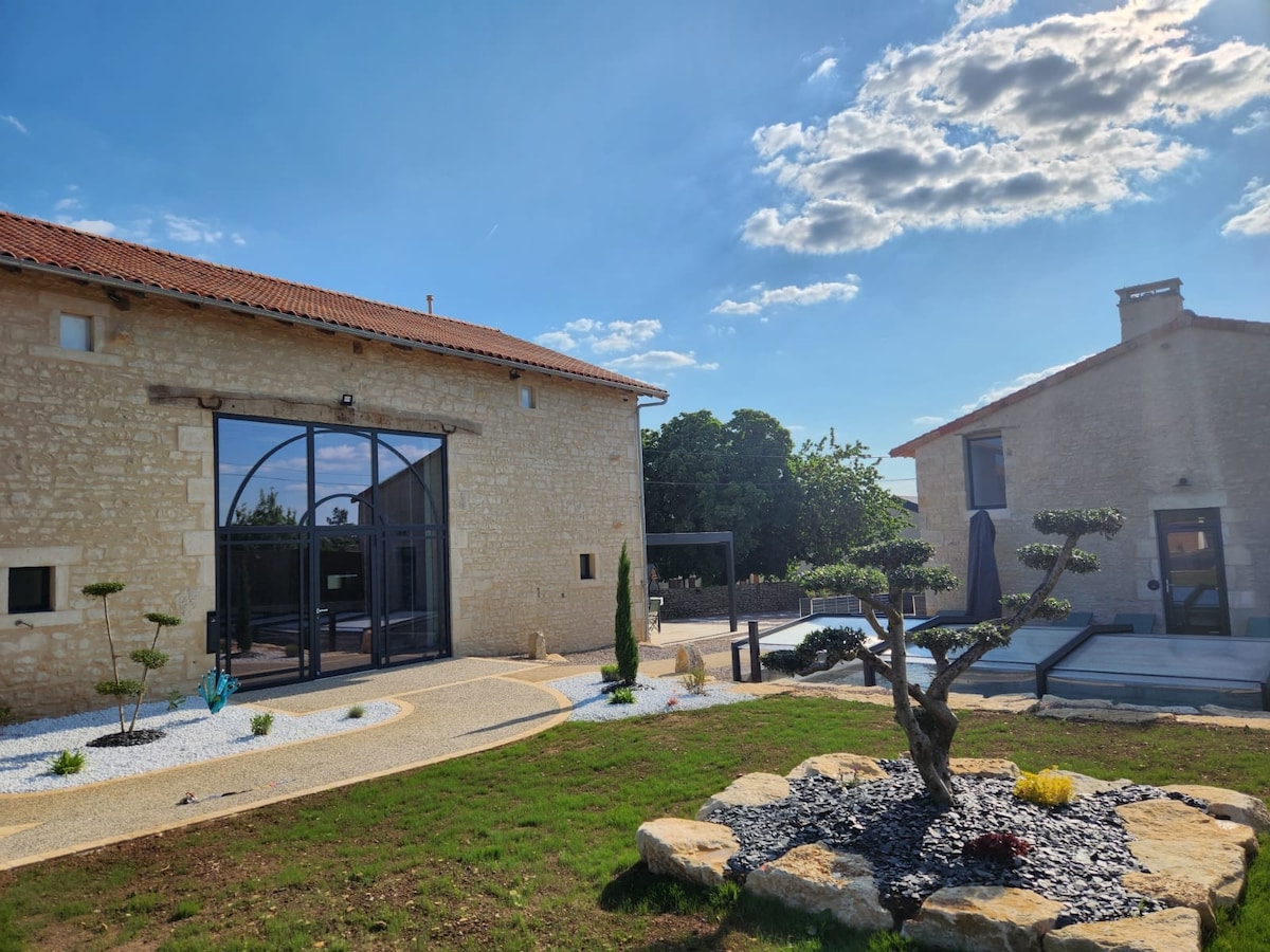 The outdoor area features a stone facade with large glass doors inviting natural light. A manicured garden surrounds the property, with decorative stones, a sculpted tree, and a seating area. A pool is visible in the background, complemented by a patio for relaxation.