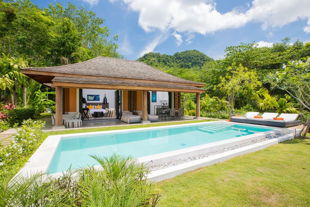 A modern bungalow is showcased with a private pool and surrounding lush greenery. The exterior features a spacious terrace, outdoor seating, and a view of the mountains in the distance. Sun loungers are positioned by the pool's edge, inviting relaxation.