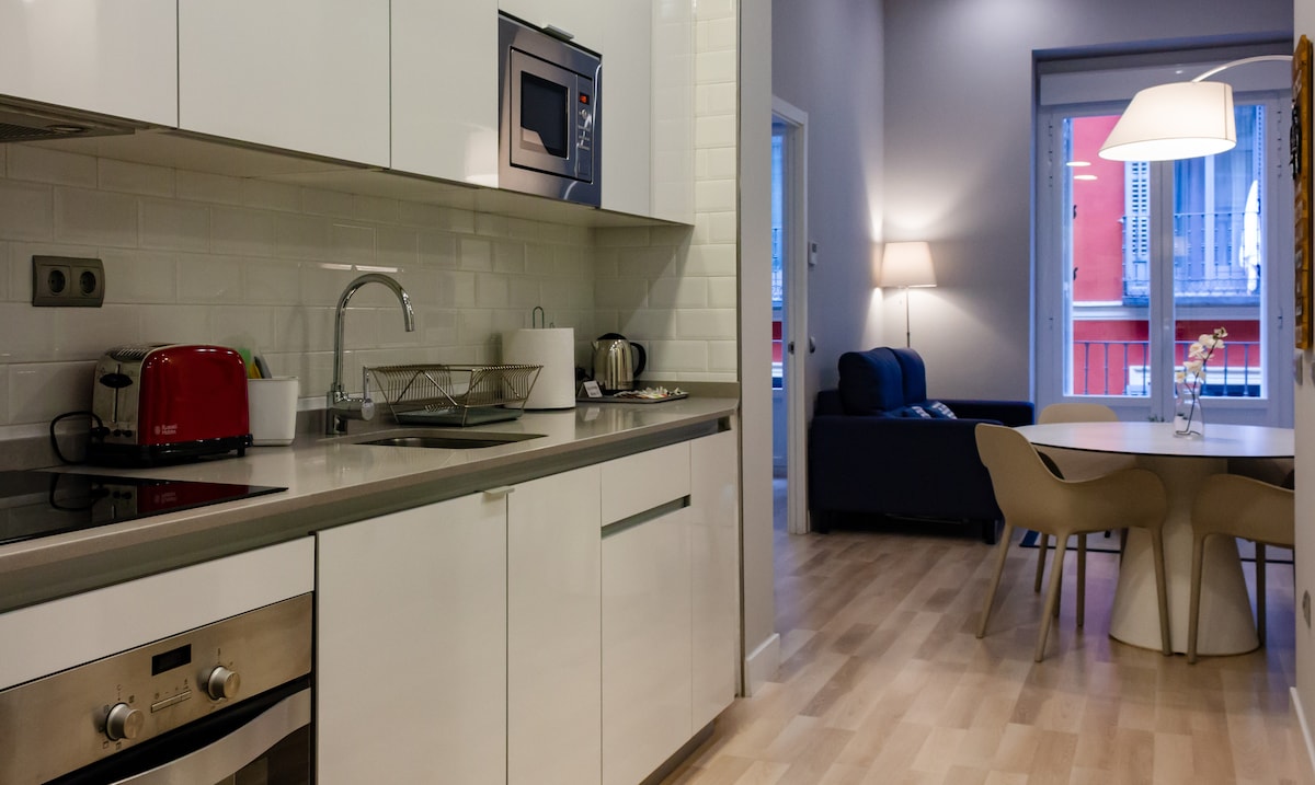 A modern kitchen features sleek white cabinetry and a countertop with a toaster, kettle, and sink. A microwave is built into the cabinetry. In the background, a cozy living area includes a sofa and a dining table, illuminated by natural light from the windows.