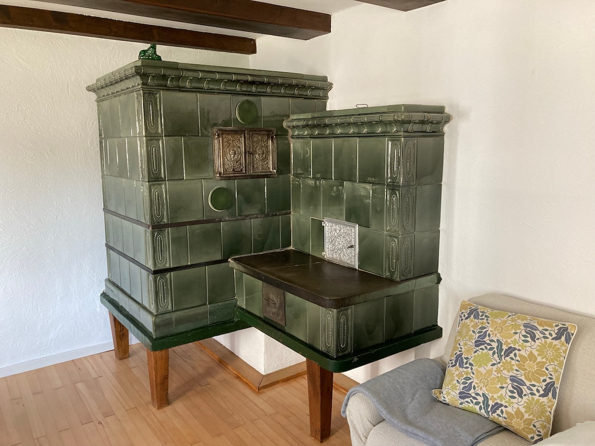 A traditional green-tiled stove features ornate detailing and is positioned against a white-walled interior. The stove is designed with a flat cooking surface, complemented by a nearby light-colored couch adorned with a patterned throw pillow.