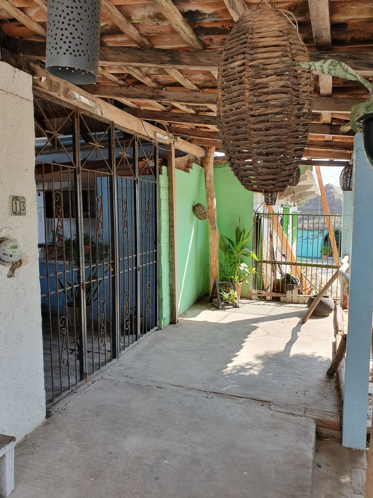 A shaded outdoor area is enclosed by a wrought iron gate, featuring a concrete floor and wooden beams overhead. Natural light filters through hanging woven light fixtures, and potted plants are positioned beside an open space leading to a bright blue room.