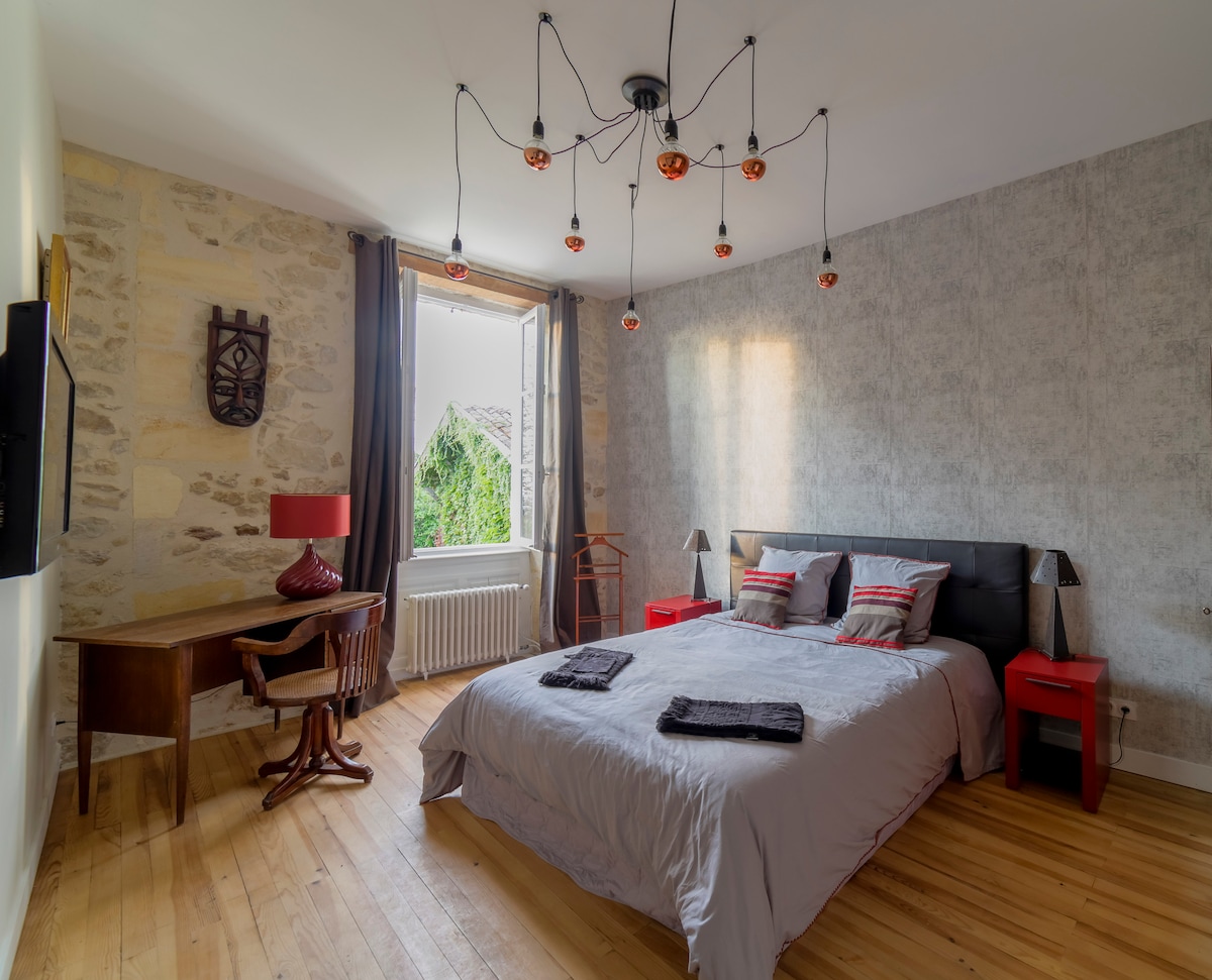 A spacious bedroom features a large bed with grey bedding, complemented by accent pillows. A wooden desk and chair are positioned near the window, which offers natural light and views of greenery. A decorative light fixture adds a modern touch to the room.
