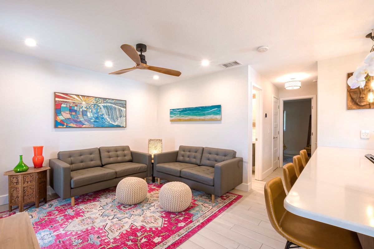 A modern living area features a pair of gray sofas and two round poufs on a vibrant area rug. Abstract wall art adds color, while a ceiling fan provides airflow. A bar counter with stools is visible in the background, contributing to a spacious feel.
