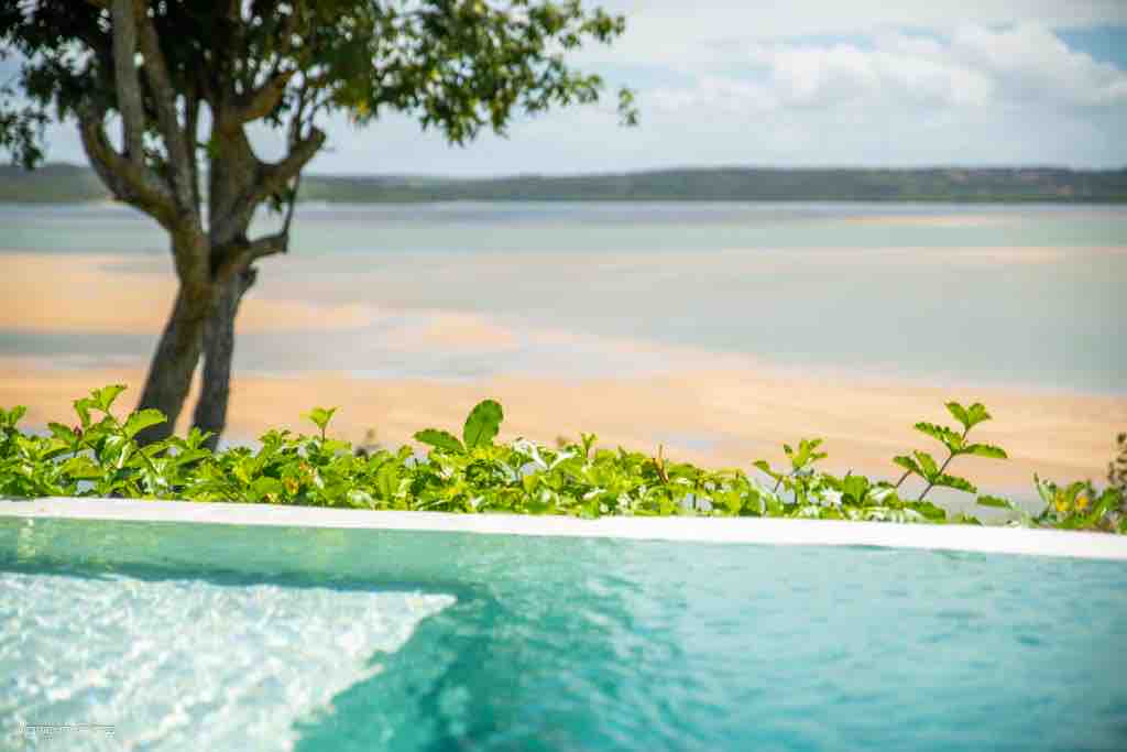 A tranquil scene is captured, featuring a private pool with crystal-clear water. Lush green foliage frames the pool, and a view of the expansive lagoon is visible in the background, showcasing gentle sandy shores under a clear sky.