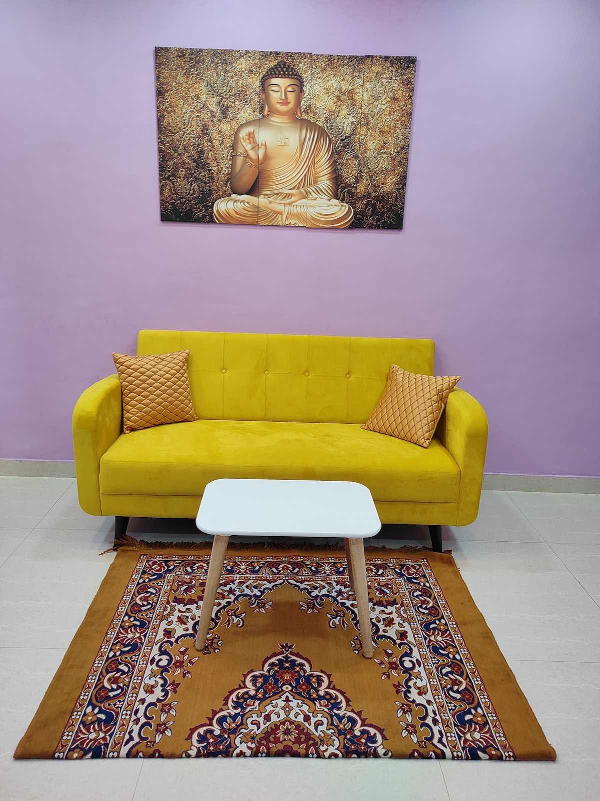 A yellow sofa with two decorative cushions is positioned in front of a white coffee table on a patterned area rug. A serene Buddha artwork is displayed on the pink wall behind the sofa, adding a calming focal point to the space.