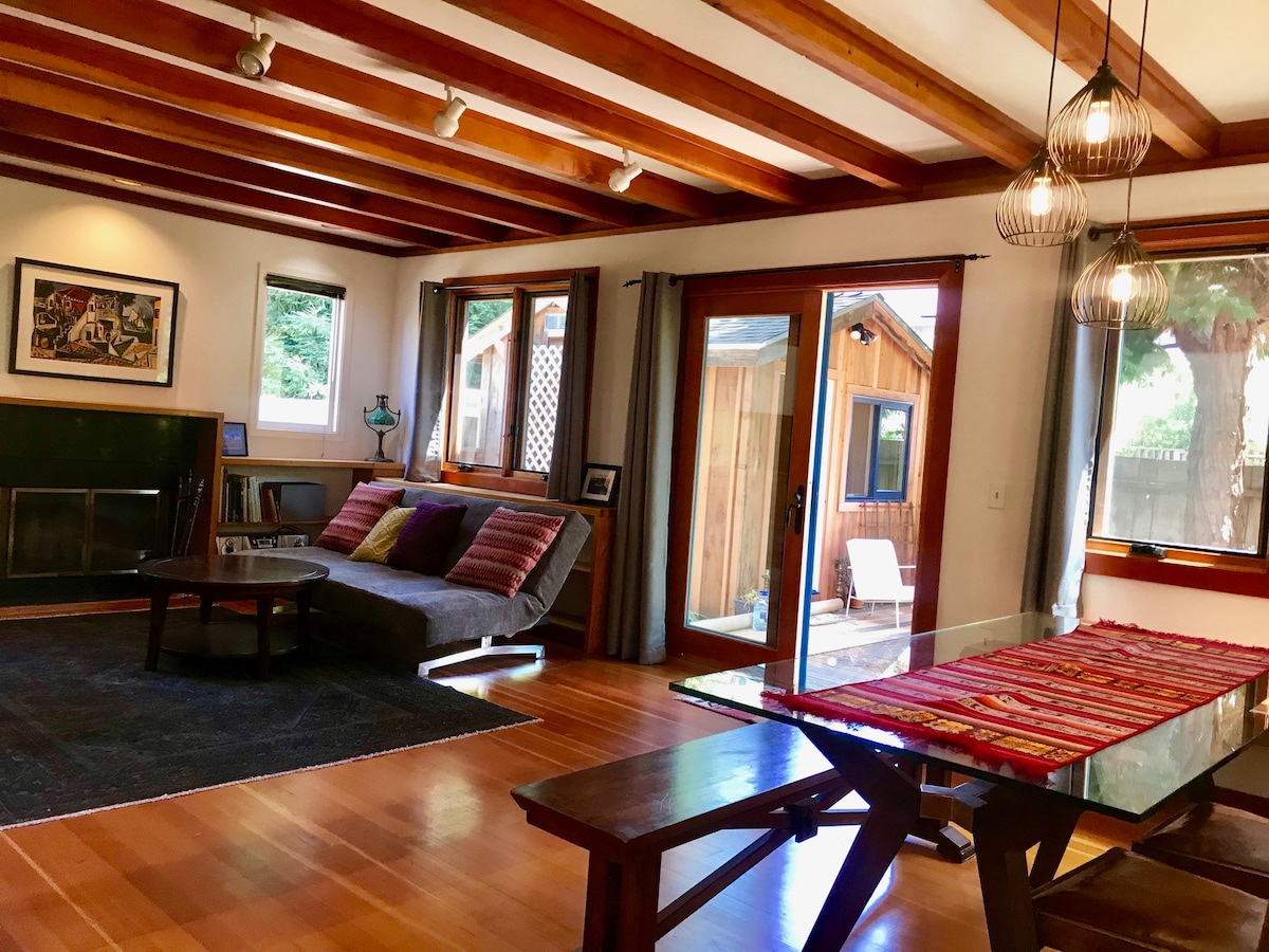 The living area is characterized by wooden beam ceilings and abundant natural light. A dark area rug anchors the space, which features a contemporary sofa adorned with cushions. A glass table is set for dining, and double doors open to the outdoor deck.