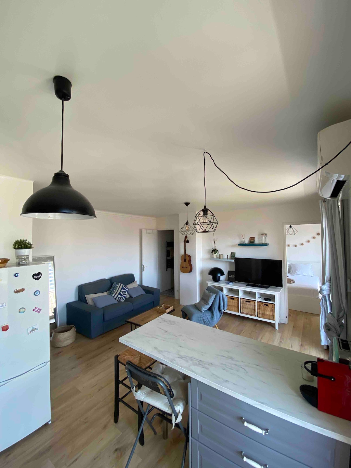 A bright living space is presented with light wood flooring and contemporary furnishings. A blue sofa is positioned against a wall, paired with a light-colored coffee table. An accent guitar decorates the space, while a sleek television sits on a low cabinet next to a minimalist white fridge.