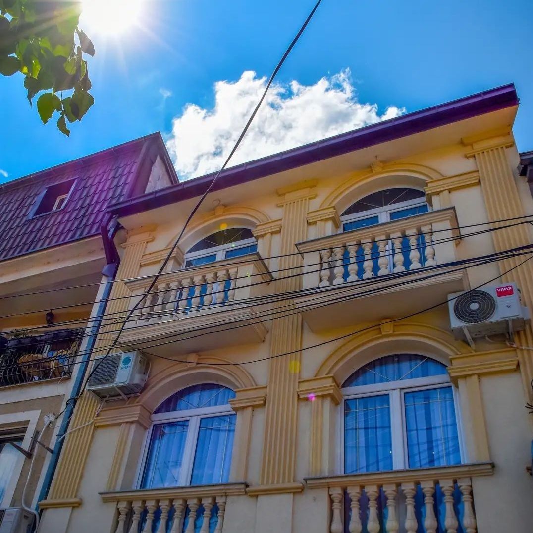 A two-story apartment building features ornate arched windows adorned with decorative railings. The façade showcases a soft yellow hue complemented by bright blue skies and scattered clouds above. Air conditioning units are visible below the windows, indicating a comfortable environment.