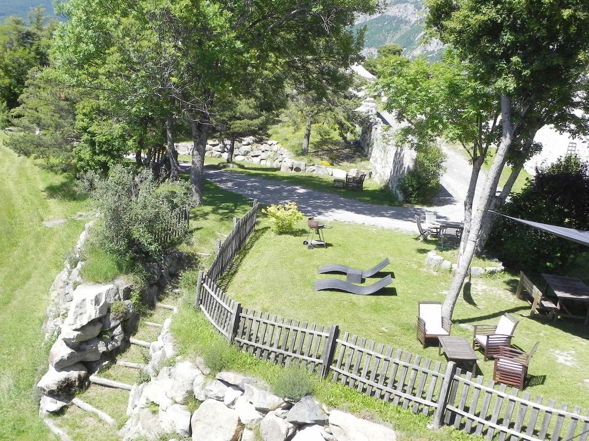 A well-maintained garden area features lush green grass and a variety of seating options, including loungers and a shaded outdoor sitting area. Stone steps lead up to the grassy space, surrounded by trees and decorative stone elements, providing a sense of tranquility.