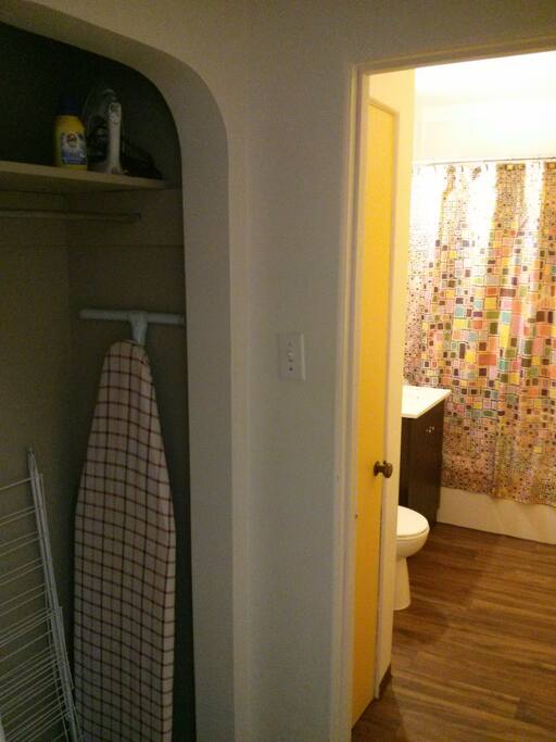 Small hall open closet with hangers, iron, ironing board, clothes drying rack, laundry detergent, shoe mat, and hanging shoe organizer.