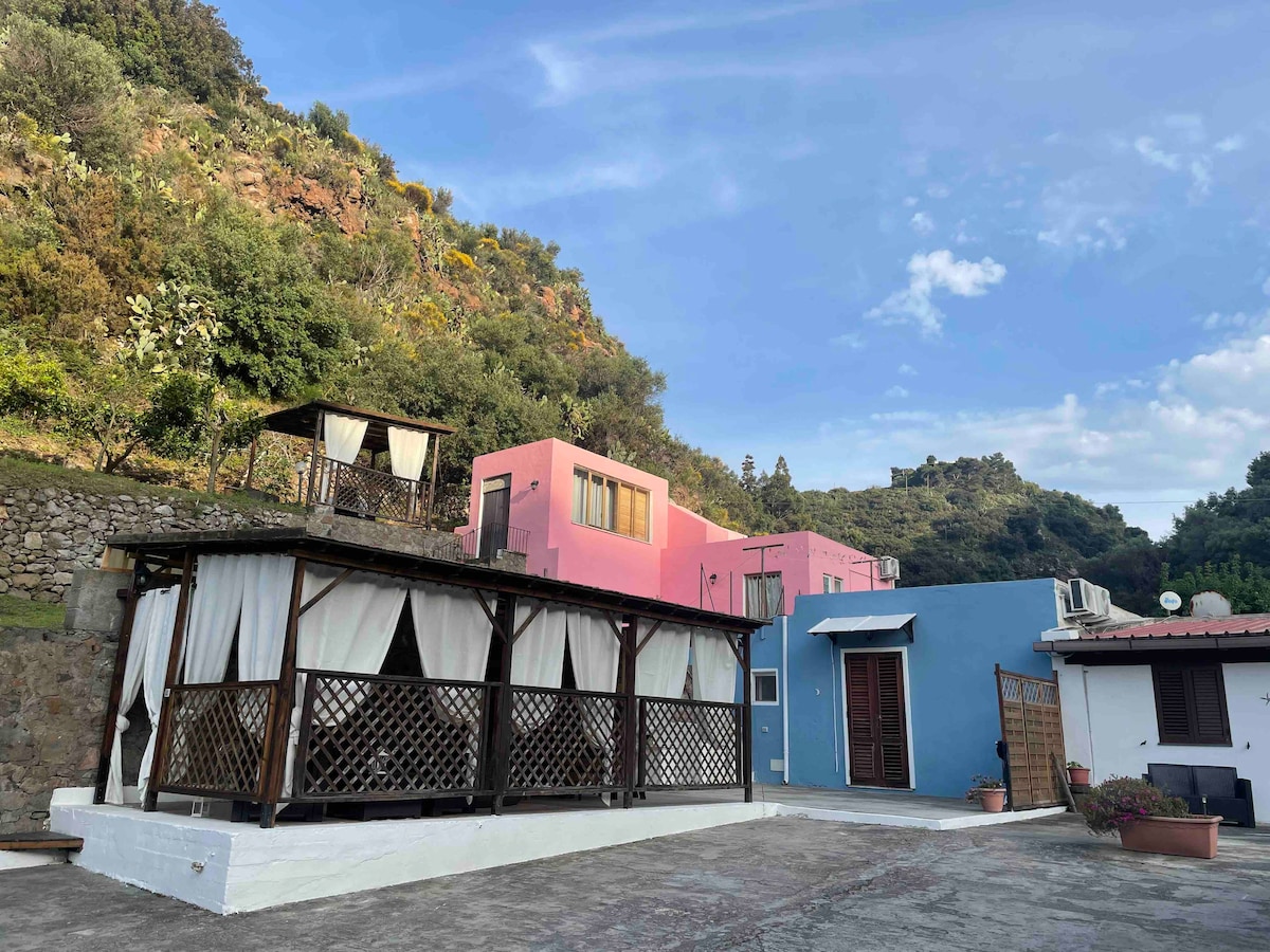 A vibrant exterior view features a spacious terrace adorned with soft curtains, surrounded by lush greenery. Colorful buildings in soft pinks and whites complement the natural landscape, while the area is illuminated by gentle daylight, creating a serene environment.