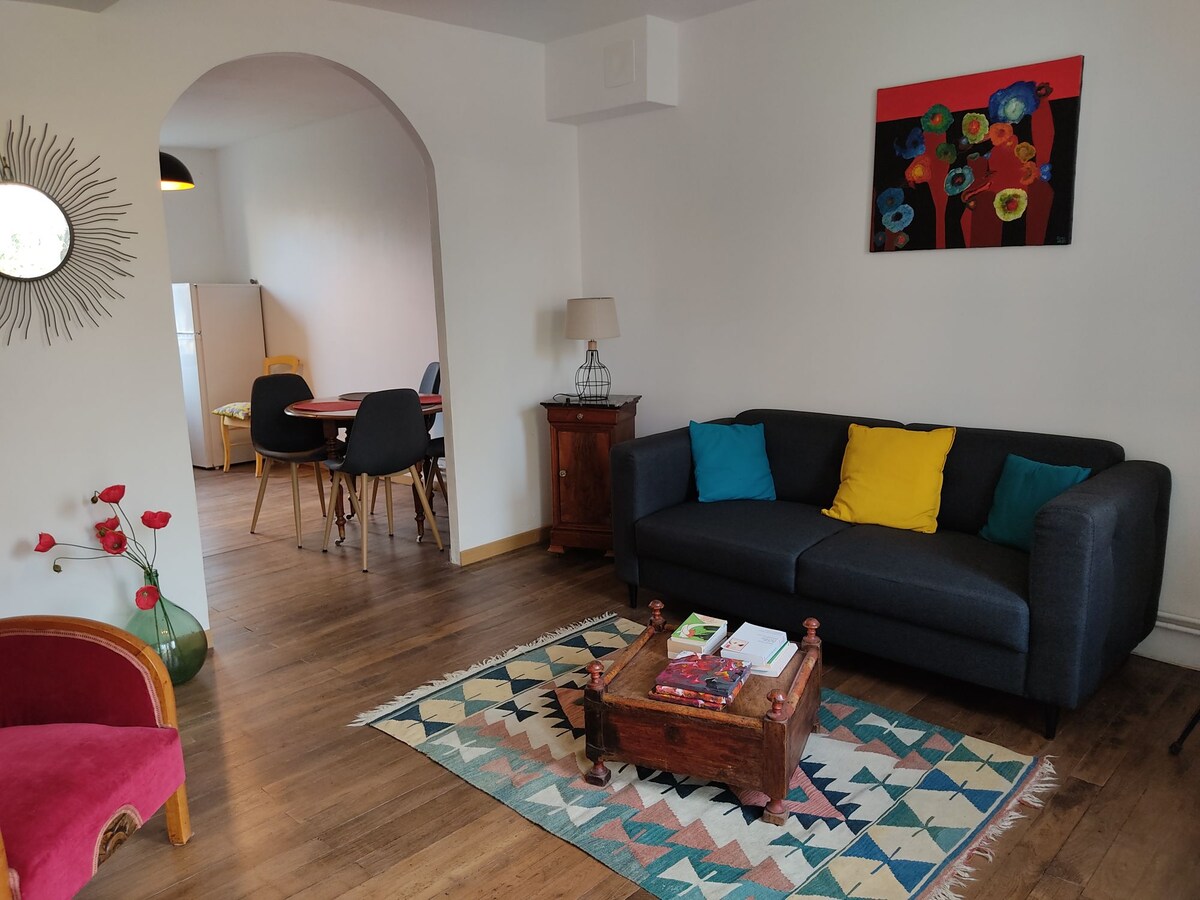 The living area features a dark sofa adorned with decorative pillows in vibrant colors. A wooden coffee table is centered on a patterned rug, accompanied by a vintage lamp. A dining space with a round table and chairs is visible in the background, creating a welcoming environment.