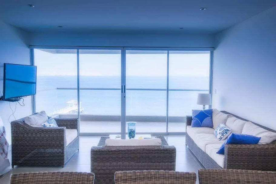 A spacious living area features large glass sliding doors that provide a stunning view of the ocean. Comfortable wicker seating is arranged around a simple coffee table, and a flat-screen TV is mounted on the wall. Soft natural light fills the space.