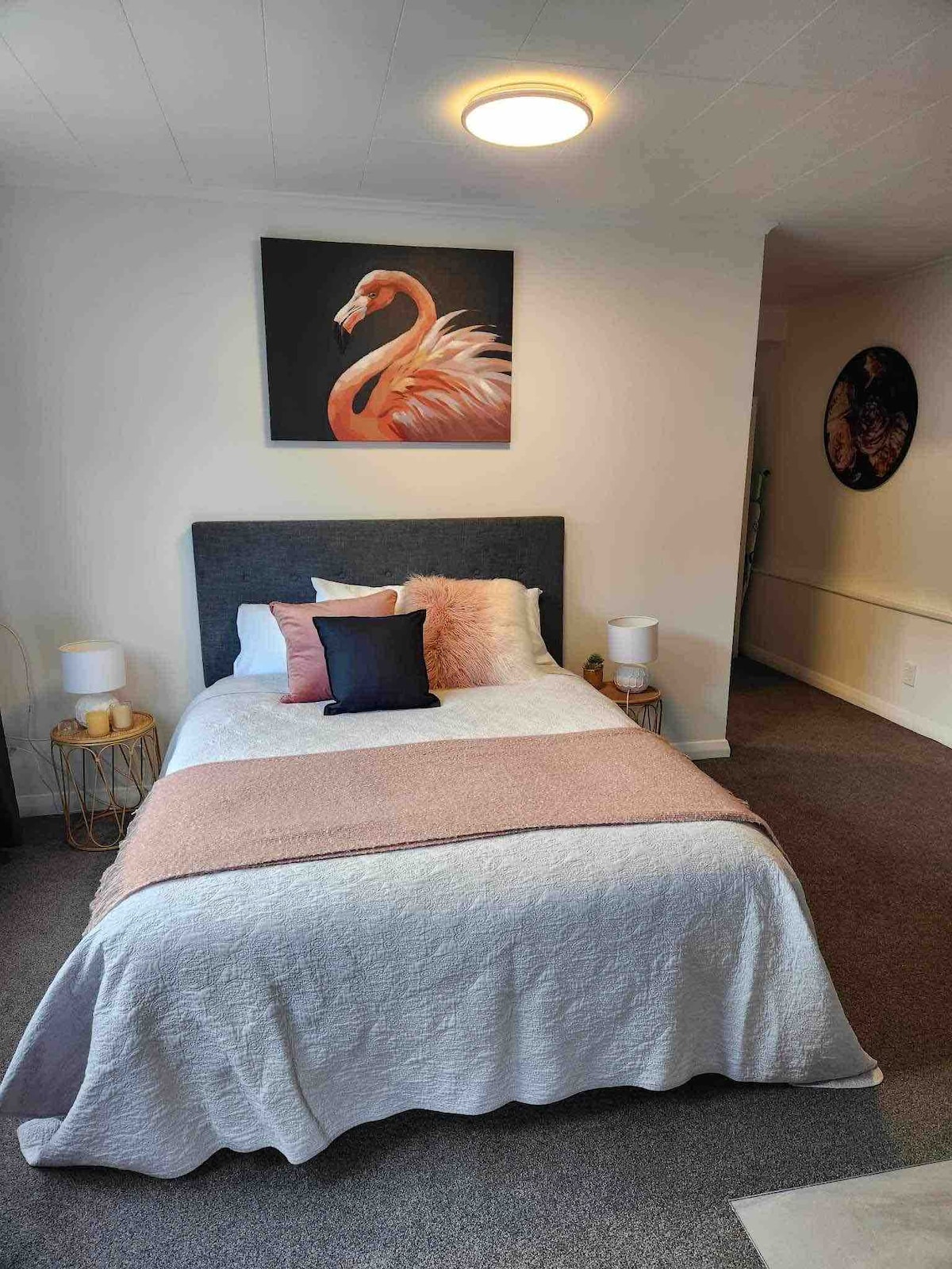 A neatly arranged sleeping area features a spacious bed with a light-colored quilt and accent pillows. A wall art piece depicting a flamingo adds a decorative touch, while a soft pink throw blanket drapes across the foot of the bed. Soft lighting is provided by bedside lamps.