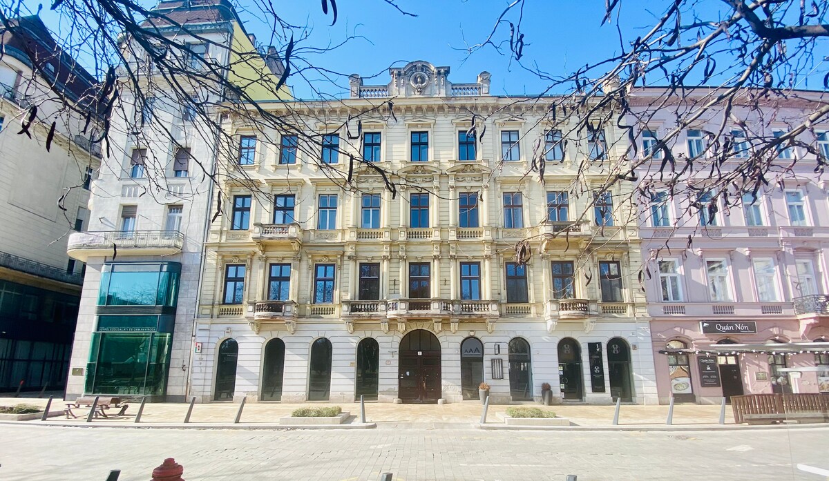 The exterior of a historic building is showcased, featuring ornate architectural details and a symmetrical façade. Balconies adorned with decorative railings line the upper floors, while large windows reflect the bright blue sky. Nearby structures illustrate the blend of modern and classic design.