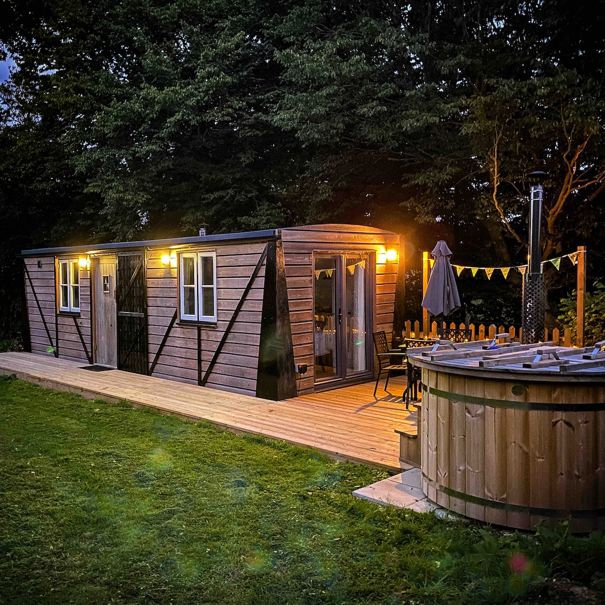 An exterior view showcases The Hoppers' Halt, with a wooden facade and warm exterior lighting. A spacious deck leads to a wood-fired hot tub, surrounded by greenery, providing a cozy setting for relaxation in the evening.