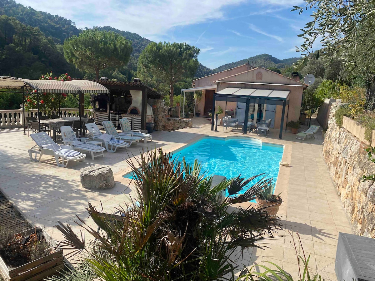 A private pool area features a sparkling blue pool surrounded by sun loungers on a tiled patio. The space is accented by lush greenery and stone walls, with a covered seating area and barbecue visible. The backdrop includes rolling hills and trees under a clear sky.