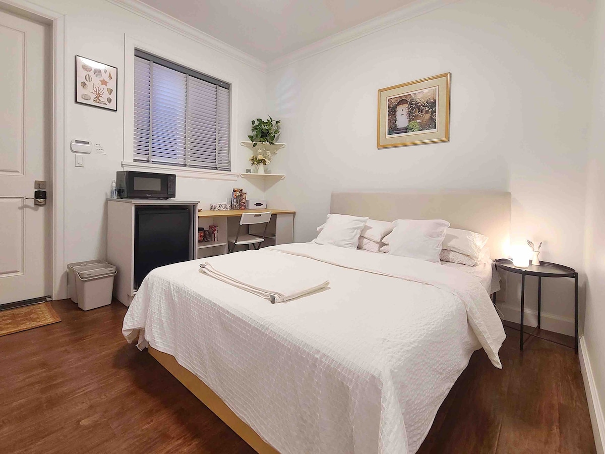 The room features a spacious bed with a light-colored quilt and multiple pillows. A small kitchenette area with a microwave is present, along with a desk and a plant for added detail. Natural light enters through a window, complemented by subtle decor elements.