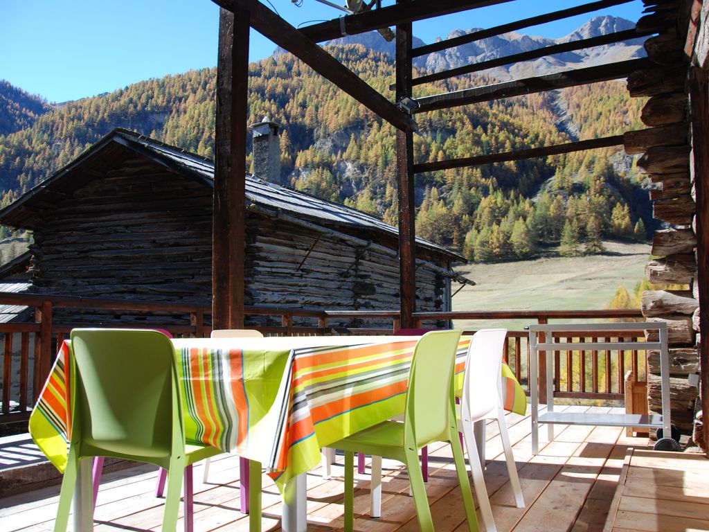 An outdoor terrace is presented, featuring a wooden table adorned with a colorful striped tablecloth. Surrounding the table are plastic chairs in various colors, offering a casual dining space. The backdrop displays mountains and trees with autumn foliage, contributing to a serene atmosphere.