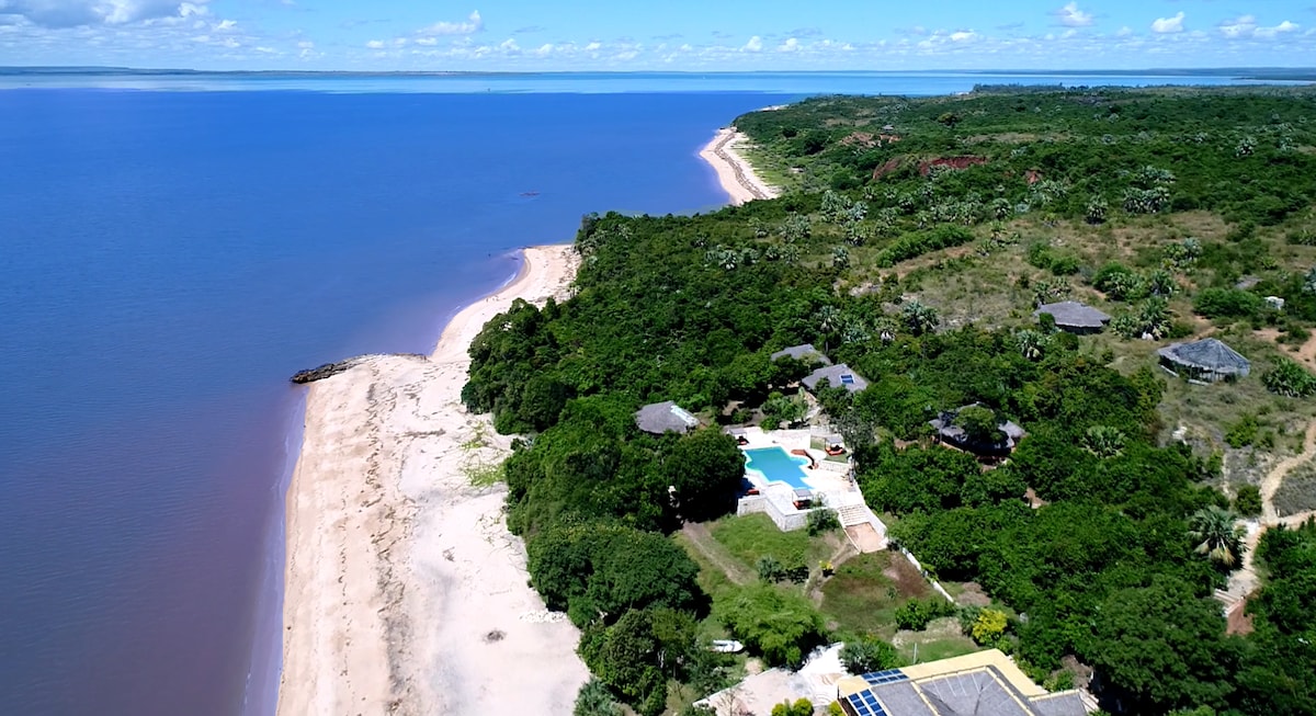 An aerial view captures the coastline stretching along the beach, bordered by lush green foliage and gentle sandy shores. The outdoor pool of the eco-lodge is visible among the trees, offering a glimpse of the natural integration with the surrounding environment.