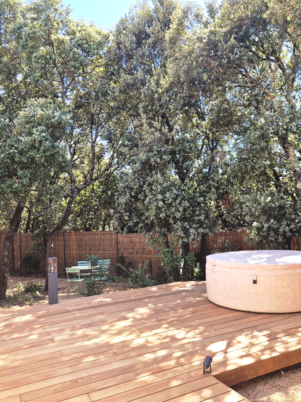 A private wooden deck features a spacious jacuzzi surrounded by lush greenery. A seating area with light blue chairs can be seen in the background, complemented by natural wood fencing that enhances the secluded atmosphere.