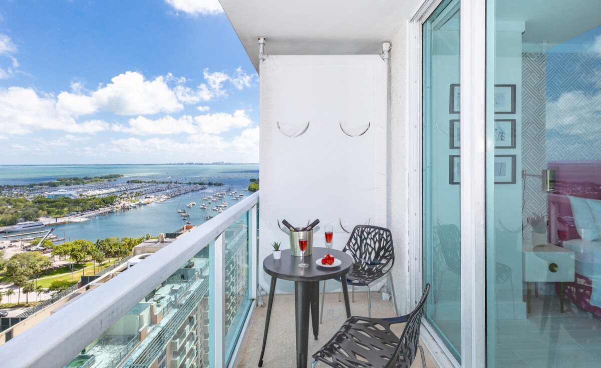 A balcony is viewed with a glass railing overlooking scenic bay and marina views. A small round table holds two drinks and a plate of strawberries, surrounded by two stylish chairs, providing a relaxing outdoor space.