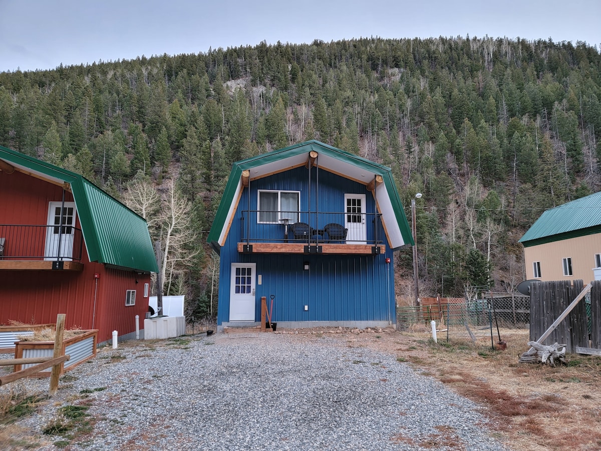The cabin features an a-frame architecture with a vibrant blue exterior contrasted by green and red neighboring buildings. The second-story balcony is furnished with chairs for outdoor seating. Surrounding trees and mountains create a natural backdrop, emphasizing the cabin's secluded location.