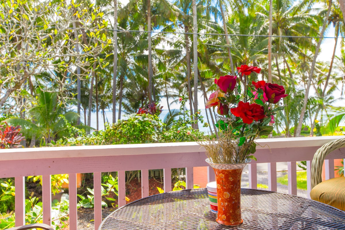 A table set on a lanai is adorned with a vase of bright red flowers. Lush greenery surrounds the area, creating a tropical atmosphere with views of the ocean visible beyond the foliage.