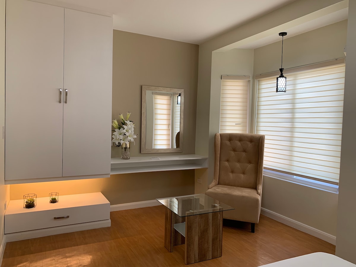A softly lit corner of the room features a plush armchair and an elegant glass coffee table. A console with a mirror sits against the wall, complemented by a work surface and two windows allowing natural light to filter through soft shades.