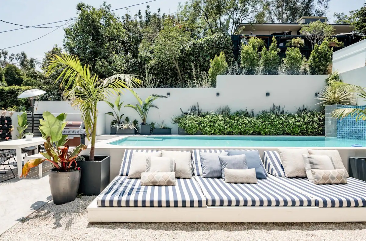 A large heated pool is complemented by a waterfall feature, surrounded by lush greenery. A built-in daybed with striped cushions provides a comfortable lounging area. Outdoor dining furniture is positioned nearby, creating an inviting space for relaxation and entertainment.