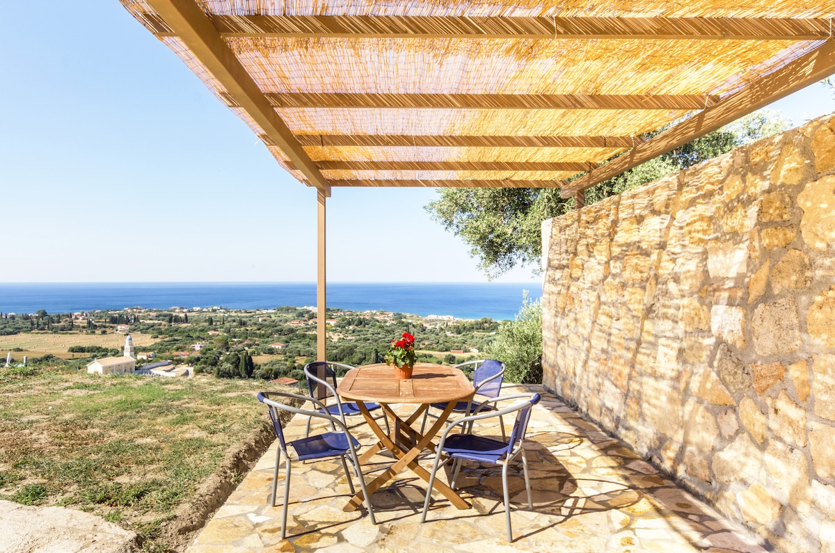 An outdoor seating area is shaded by a straw-covered pergola, featuring a round wooden table surrounded by four blue chairs. The paved stone floor extends to lush greenery, offering unobstructed views of the coastline and the sea in the distance.