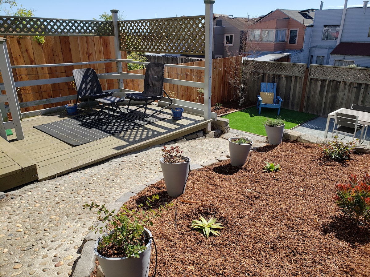An outdoor seating area features two lounge chairs set on a wooden deck, surrounded by landscaped gravel and potted plants. A fence creates privacy, while a small patch of artificial grass is visible in the background, enhancing the inviting space for relaxation.