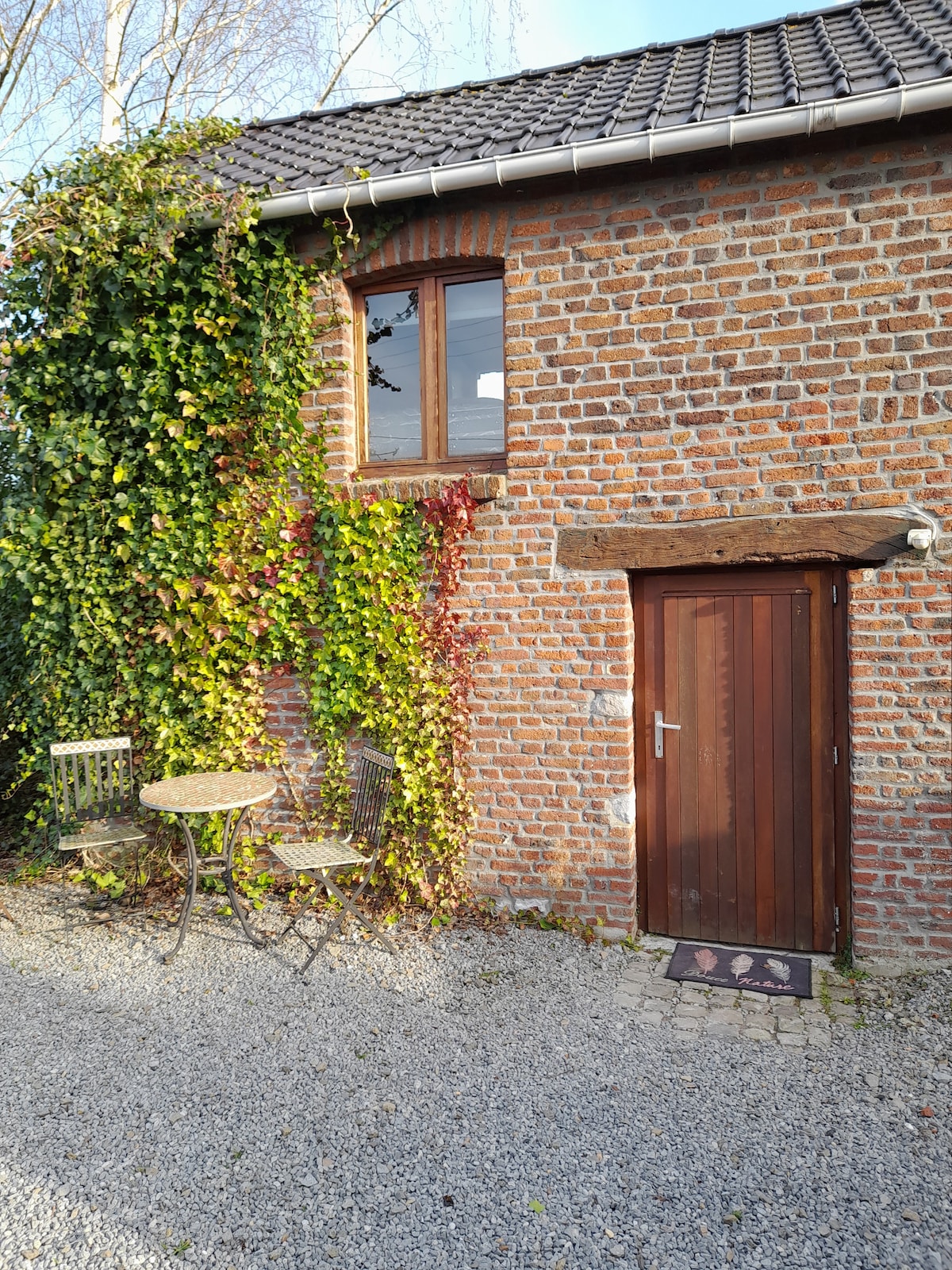 The exterior of the micro house features a rustic brick facade with a wooden door. Ivy clings to the wall, adding greenery, while a small bistro table and two chairs are positioned on gravel. A single window above allows natural light to enter.
