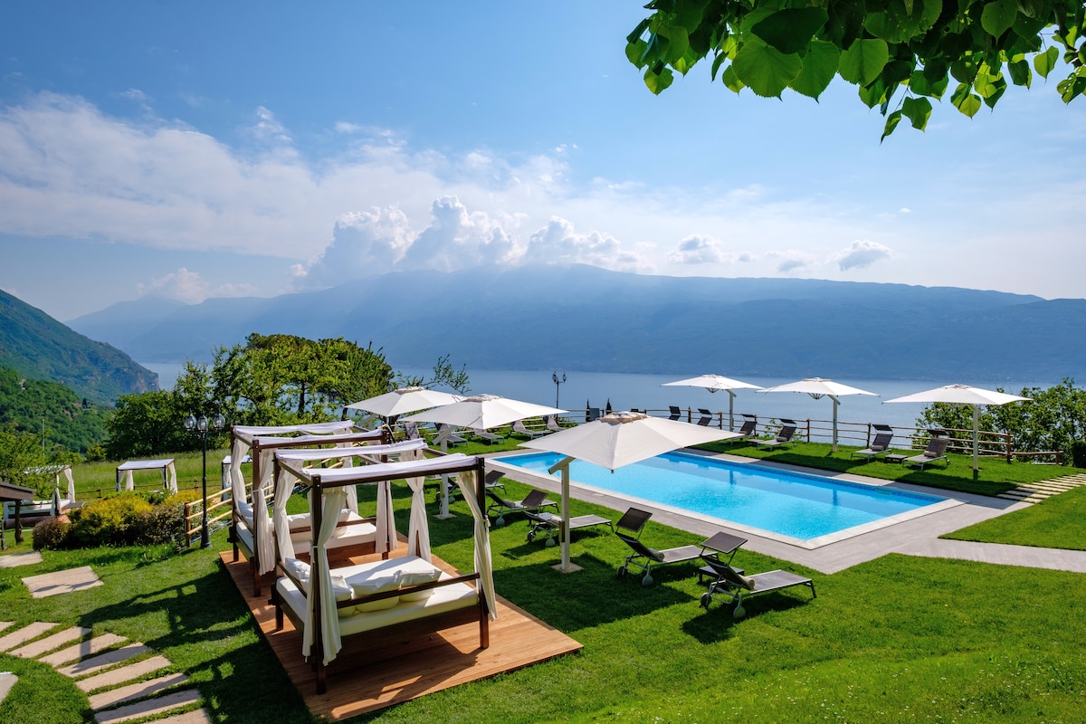 An expansive outdoor area features a clear swimming pool surrounded by sun loungers and shaded seating areas. The lush green landscape offers views of the lake and mountains under a bright sky, creating a serene and tranquil setting.