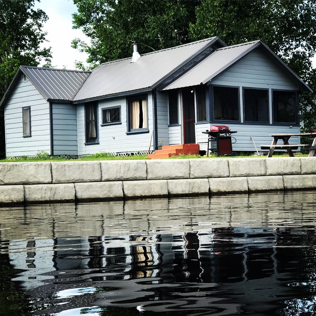 A charming single-story cabin is seen from the water's edge, featuring a metal roof and large screened-in porch. Reflective water surfaces highlight the cabin's form and surrounding greenery. A picnic table and gas grill are positioned on the patio, enhancing the outdoor living space.
