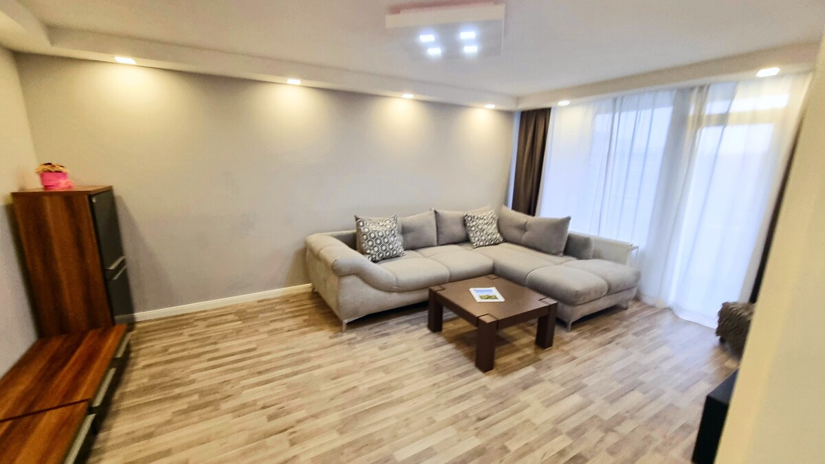 The living area features a spacious sectional sofa arranged around a wooden coffee table. Natural light enters through large, sheer-curtained windows, illuminating the light wood flooring. An entertainment unit is positioned against one wall, adding functionality to the cozy space.