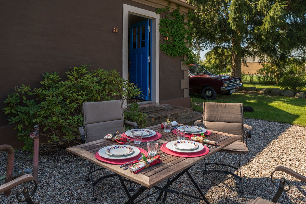An outdoor dining area is set up with a wooden table, featuring four plates and glassware. Two gray chairs are positioned at the table, with a bright blue door visible in the background, and a classic car parked nearby. The surrounding greenery adds a natural touch.