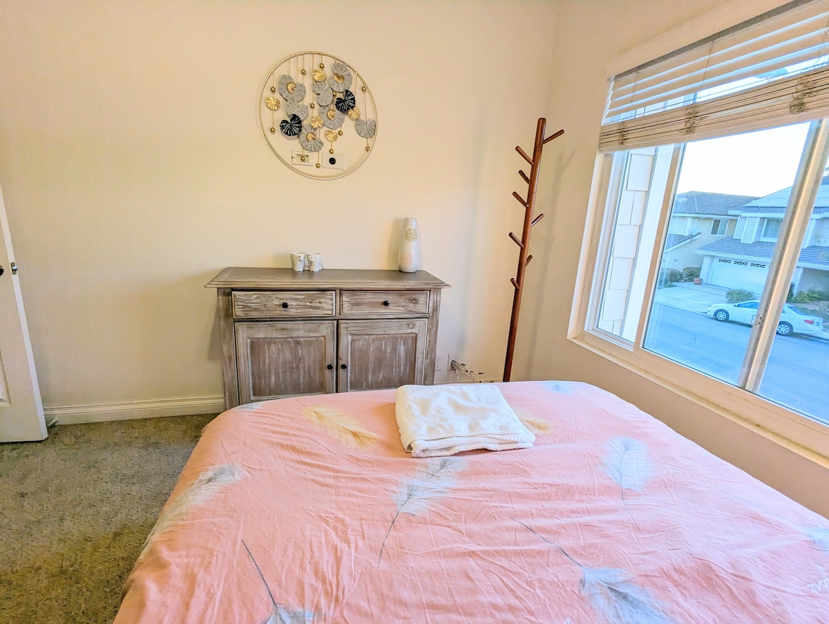 A cozy bedroom features a spacious bed with a soft, pastel pink coverlet adorned with leaf patterns. A wooden cabinet sits against the wall, accompanied by a water carafe and two glasses. Natural light floods the room through a large window adorned with simple blinds.