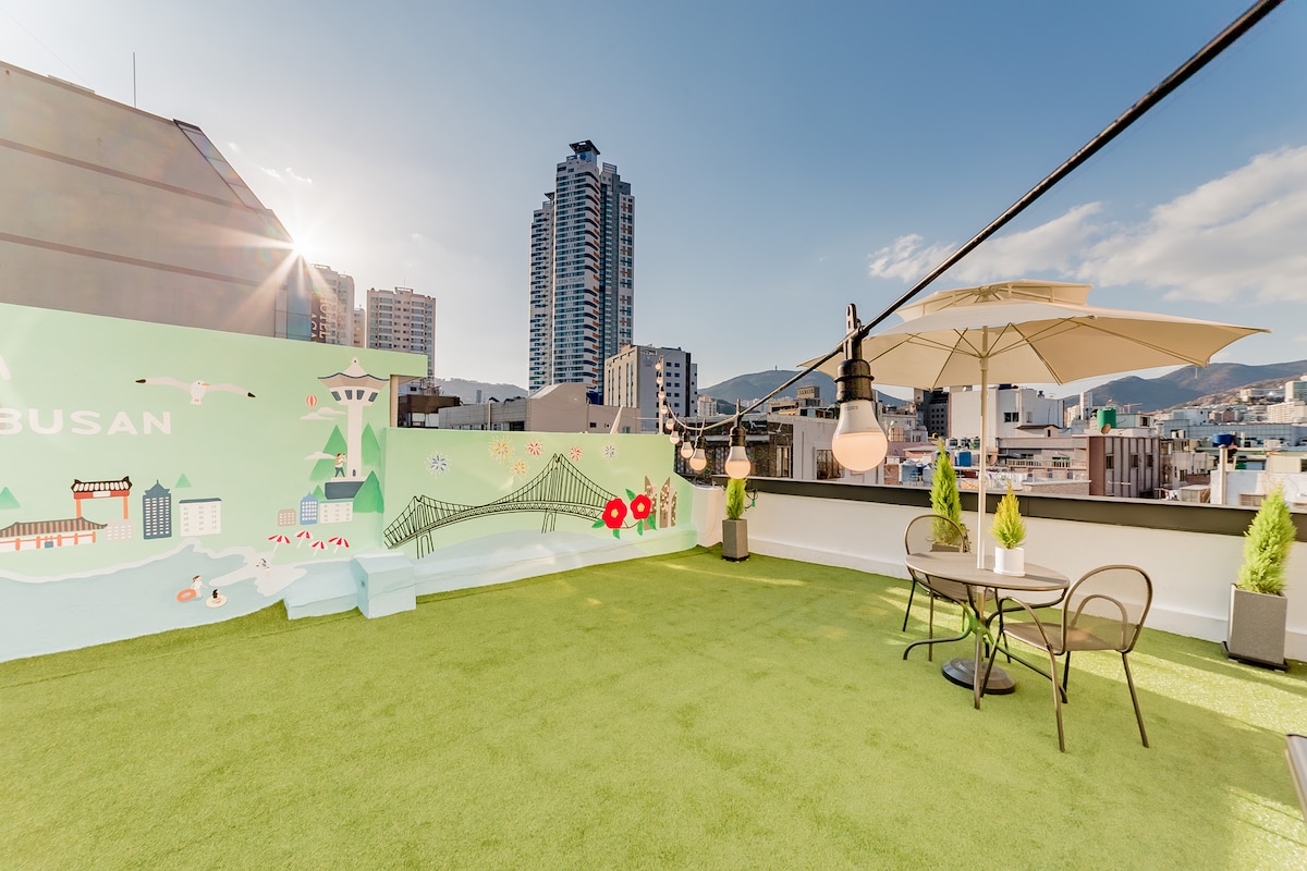 A rooftop area features green artificial grass, providing a relaxed setting. A round table with chairs is present, accompanied by a large umbrella for shade. Scenic views of the city skyline and a decorative mural enhance the inviting ambiance of this outdoor space.