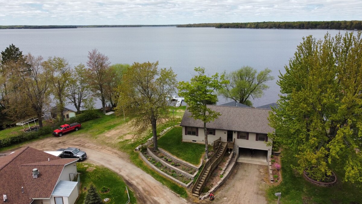 An aerial view captures a waterfront home surrounded by trees and a sandy pathway leading to Pelican Lake. The lake reflects the cloudy sky, while the lush greenery and landscaped steps enhance the setting. Nearby vehicles and neighboring homes are subtly visible.