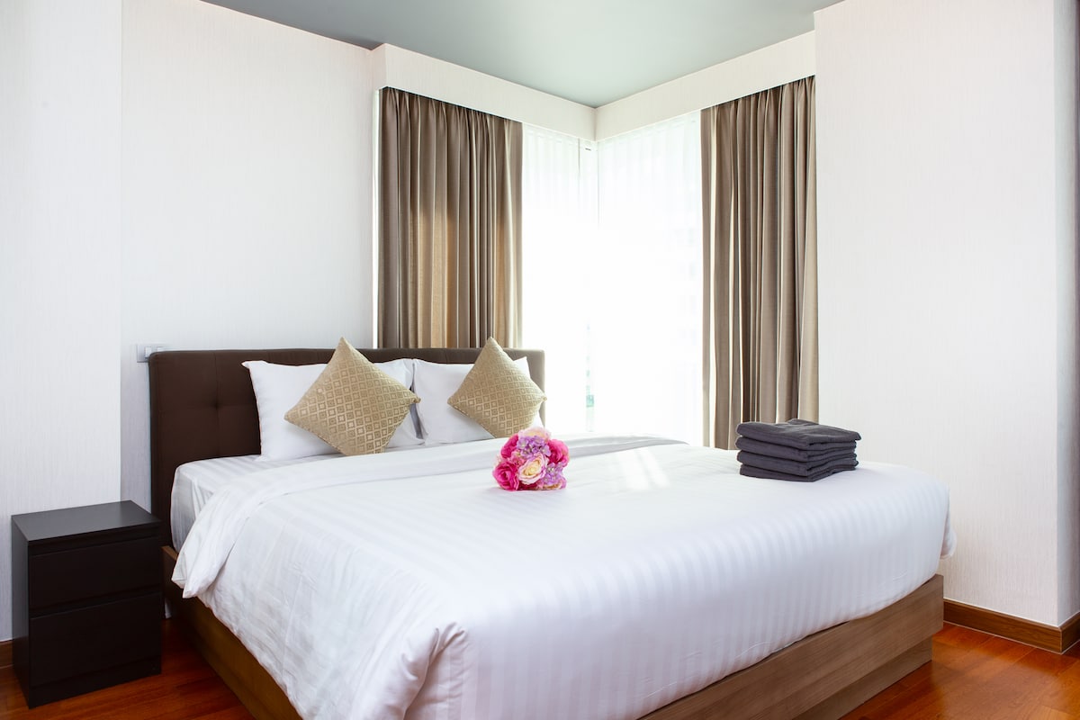 A spacious bedroom is adorned with a king-size bed featuring crisp white bedding and decorative cushions. Soft curtains frame large windows, allowing natural light to fill the room. A nightstand and neatly arranged towels are visible, contributing to the well-organized space.